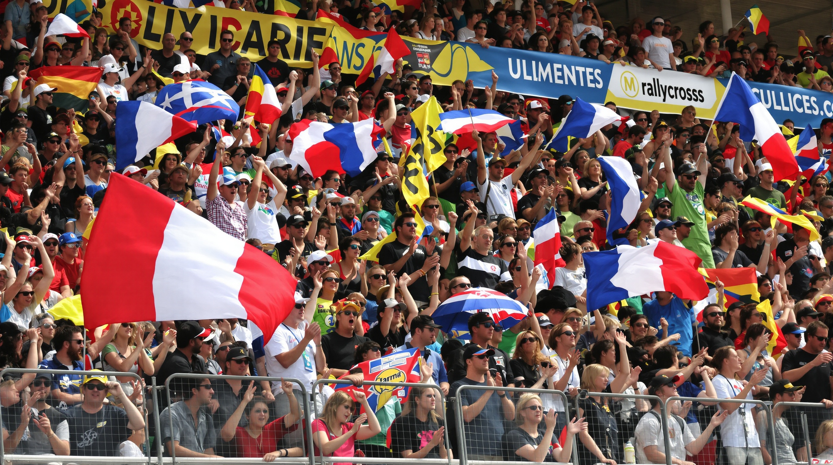 Scène de foule enthousiaste sur le circuit Maurice Forget à Mayenne, un samedi ou dimanche de rallycross 2025, drapeaux, public excité, gros plan sur la tribune en liesse et ambiance d'attente avant le départ.